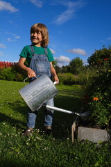 Summertime, sweet happy boy helping grandmother in the country. Full length portrait of child in the t-shirt and denim jumpsuit with the watering can in his hands watering flowers in the garden