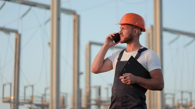 Busy caucasian man in helmet and uniform talking on smartphone and holding clipboard while standing among power station. Concept of people, electricity and technology.