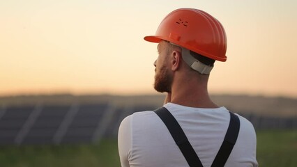 Back view of male technician in white helmet staning during evening time among field with solar panels. Caucasian man checking and eximing production of green energy.
