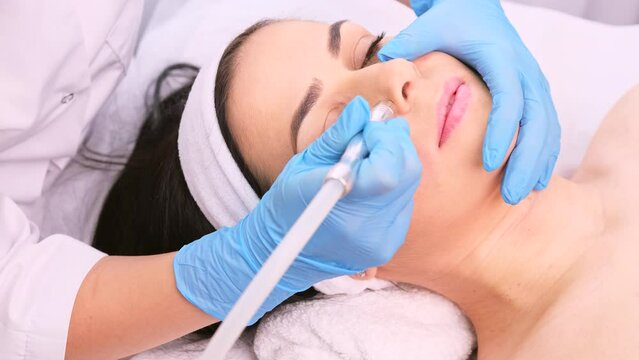 Cosmetologist in blue gloves making rejuvenation procedure with a vacuum apparatus massage microdermabrasion instrument to a brunette woman.