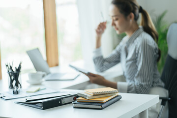 Obraz premium Side view. Young asian business woman sitting at table and taking notes in notebook.On table is laptop, smartphone and cup of coffee. Student learning online. Blogger.