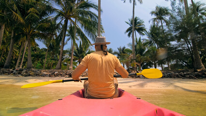 Young man with sunglasses and hat rows pink plastic canoe along sea against green hilly islands with wild jungles. Traveling to tropical countries. Strong guy is sailing on kayak in ocean, back view.