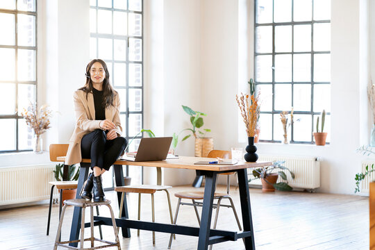 Businesswoman wearing headset sitting by laptop on table at home office - Powered by Adobe
