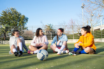 Portrait of young girls gathering on football field. Four beautiful girls in sportswear sitting on green field resting and discussing game before playing. Healthy lifestyle and sport activity concept