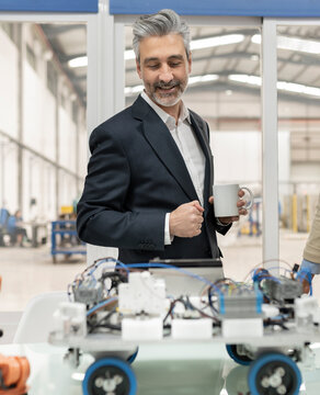Smiling businessman holding coffee cup looking at robotic vehicle model standing at desk