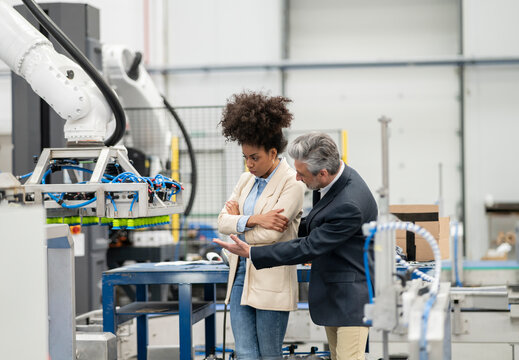 Engineer explaining robotic arm to developer standing with arms crossed in factory
