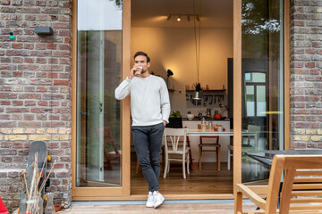 Young man drinking coffee standing with hand in pocket leaning on doorway