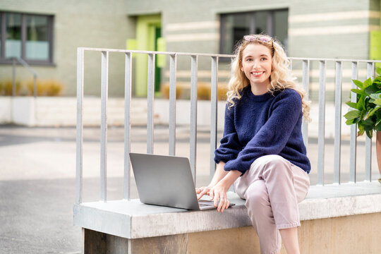 Happy young blond woman sitting with laptop on wall ledger