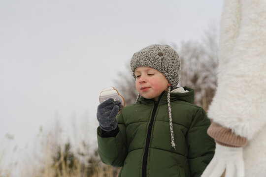 Boy wearing knit hat eating cookie standing by mother in winter