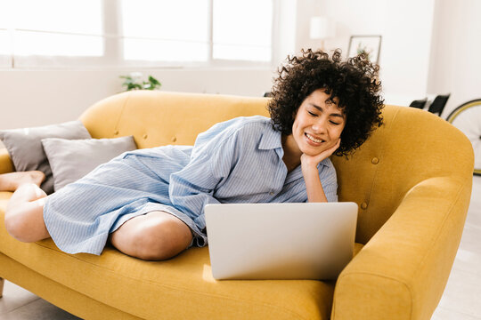 Smiling young woman using laptop lying on sofa in living room at home