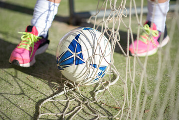 Fototapeta na wymiar Close-up of football ball lying on green field. Clean white and blue ball lying on net of football goal and female player standing near preparing to kick. Sport activity and healthy lifestyle concept