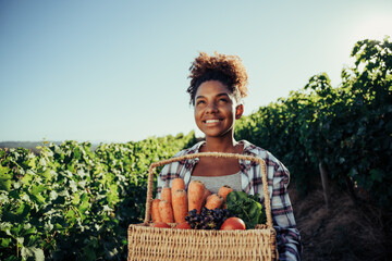 Mixed race female walking through vineyards smiling holding large basket of vegetbales 