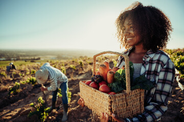 Mixed race female farmer holding vegetable basket while male worker harvests crops 