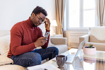 African astonished man sitting on sofa holds cellphone read e-mail sms feels shocked received terrified news, guy looks at online calendar forgot missed important meeting, phone problems concept..