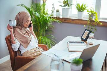 Smiling Muslim woman talking on the phone, studying online with a Skype teacher, happy young Muslim...
