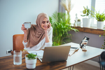 A positive Arab woman in a hijab drinks coffee, has an online meeting on a laptop at home. A Muslim business woman in a headscarf participates in a webinar, working remotely.