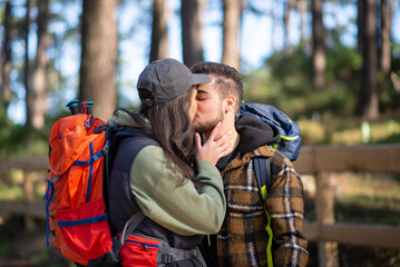 Obraz premium Affectionate couple of young hikers. Caucasian man with beard and woman in cap with big backpacks kissing in forest. Hobby, nature, love concept