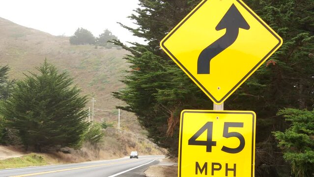 Speed limit sign, yellow warning or caution about curves, turns or winding road ahead. Pacific coast highway trip, cabrillo highway along ocean coast. Cars driving on asphalt. California Big Sur, USA.