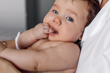 Portrait of naked baby lying in mothers arms at home, blurred background. Infant child under parental protection in bedroom closeup, explore world. Home shooting, free copy space. Childcare concept.