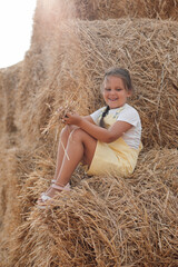 Naklejka na ściany i meble Shining beautiful female child on hayrick smiling looking down and having some hay in hands wearing sundress. Having fun away from city on field full of golden hay.