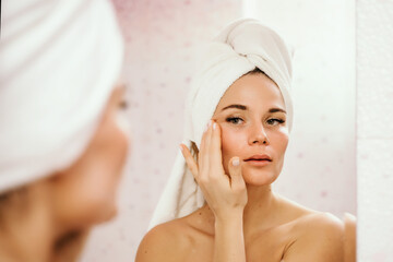 Young beautiful woman using face cream moisturizing lotion after bath. Pretty attractive girl in a towel on her head stands in front of a mirror in a home bathroom. Daily hygiene and skin care