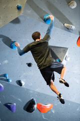 Back view of young guy exercising climbing in gym. Caucasian athlete doing bouldering holding...