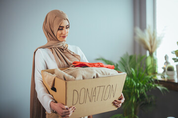 Muslim woman with donation box at home. Donation box for poor with clothing in female hands. Woman...