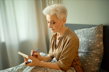 Indoor picture of serious concentrated senior female with grey short hair noting down her plans,...