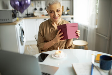 Fototapeta na wymiar Attractive senior grey haired woman in light brown dress sitting at kitchen table with present box in hand, having online celebration via laptop web camera, smart phone and cupcake on table