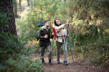 Active young hikers walking in forest. Caucasian man and woman in casual clothes with big backpacks trekking, pointing, discussing. Hobby, nature, love concept