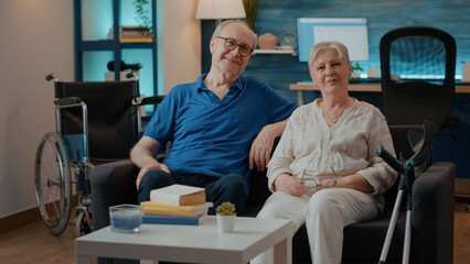 Portrait of retired couple with disability sitting on couch, looking at camera. Elder man and woman next to crutches and wheelchair for accessibility and mobility, enjoying free time.