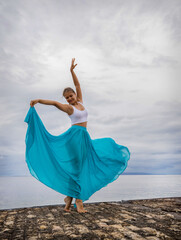 Beautiful woman practicing ballet pose. Young ballet dancer wearing long blue skirt and posing. Outdoor exercise. Slim body. Cloudy sky background. Copy space. Bali