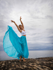 Caucasian woman practicing ballet pose. Young ballet dancer wearing long blue skirt and posing. Outdoor exercise. Slim body. Cloudy sky background. Copy space. Bali