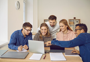 Creative university students sitting at library desk, looking at modern laptop computer, showing, watching, discussing videos, making business, entrepreneurship, marketing group project presentation