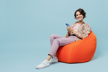 Full size young smiling cheerful happy woman 20s in brown shirt sit in bag chair hold use mobile...