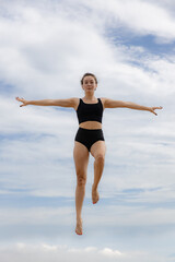 Fototapeta na wymiar Beautiful young woman jumping over cloudy blue sky. Caucasian woman wearing black sportswear. Fitness, wellness concept. Outdoor activity. Copy space. Sky background. Bali