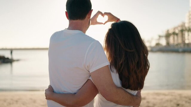 Man and woman couple hugging each other doing heart gesture with hands at beach