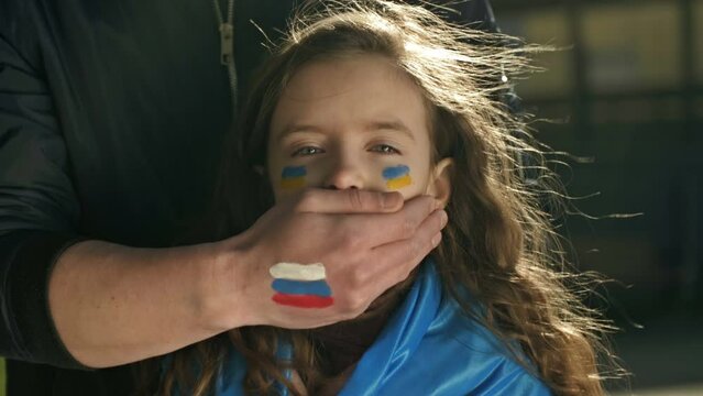 Man's hand with the image of the Russian flag roughly closes the mouth of a little girl with the Ukrainian flag. Children's tears of war. Freedom for Ukraine. Russian aggression.