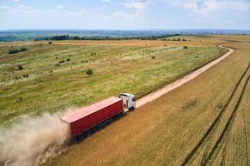 Aerial view of cargo truck driving on dirt road between agricultural wheat fields making lot of...
