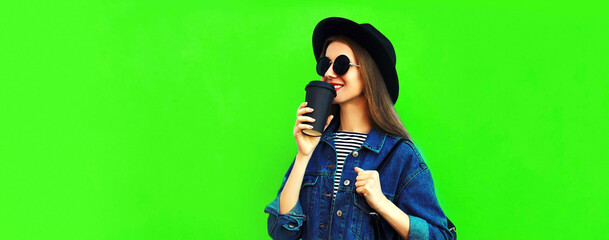 Portrait of modern young woman drinking coffee wearing black round hat, denim jacket on green background, blank copy space for advertising text