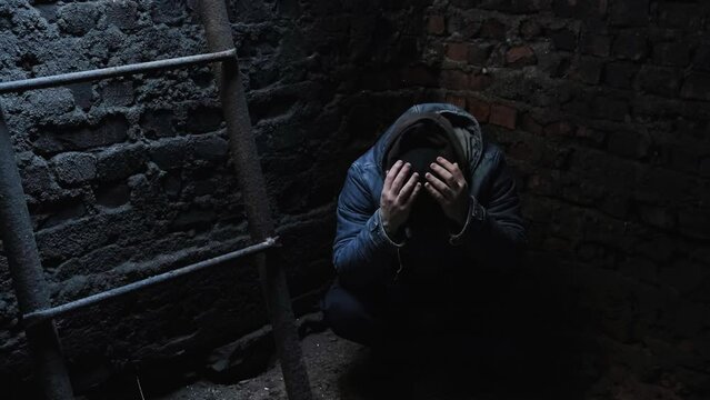 A man hides in a bomb shelter covering their head with hands. The human sits on the floor in dungeon during shelling. Terrible conditions. Fear, danger, hunger and cold. Concept of wartime. Life save.