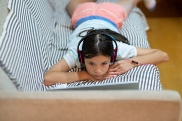 Young girl, child of mixed ethnicity laying on the sofa listening to music on headphones and using a laptop.
