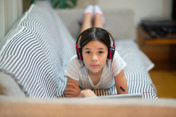 Cute young girl of mixed ethnicity, laying on the sofa listening to music on headphones and using a laptop.