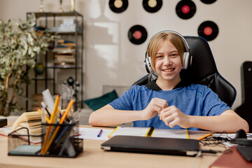 Smiling boy with wireless headphones hangs out in youthful boy's room. The teenager is sitting in comfortable armchair by desk on which are spread books, notebooks, laptop for online learning.