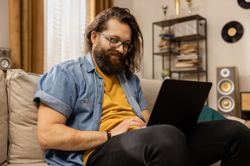 A bearded man is browsing through a gallery of vacation photos on his laptop while sitting on the couch. A handsome lumberjack-type boy uses his computer in his free time.