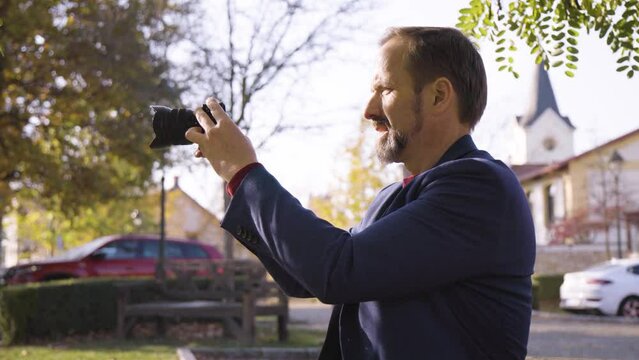 A middle-aged handsome Caucasian man takes pictures with a camera as he sits on a bench in a quaint town in fall