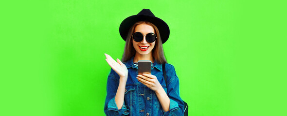 Portrait of stylish happy smiling young woman model with smartphone wearing black round hat, denim jacket on green background