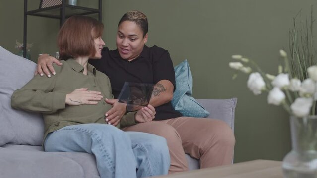 Medium slowmo of two interracial enamored women waiting for baby sitting on sofa at home with their hands on belly and looking at baby ultrasound image