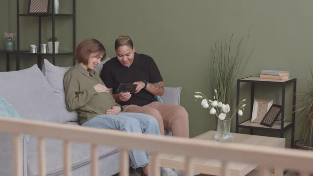 Slowmo of loving interracial lesbian couple looking at baby ultrasound image sitting on couch at home. Biracial woman kissing her beloved female partner in head