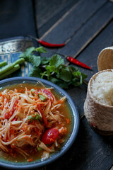 Spicy papaya salad served in a blue dish with sticky rice in a bucket and long beans, dried chilli and vegetables.